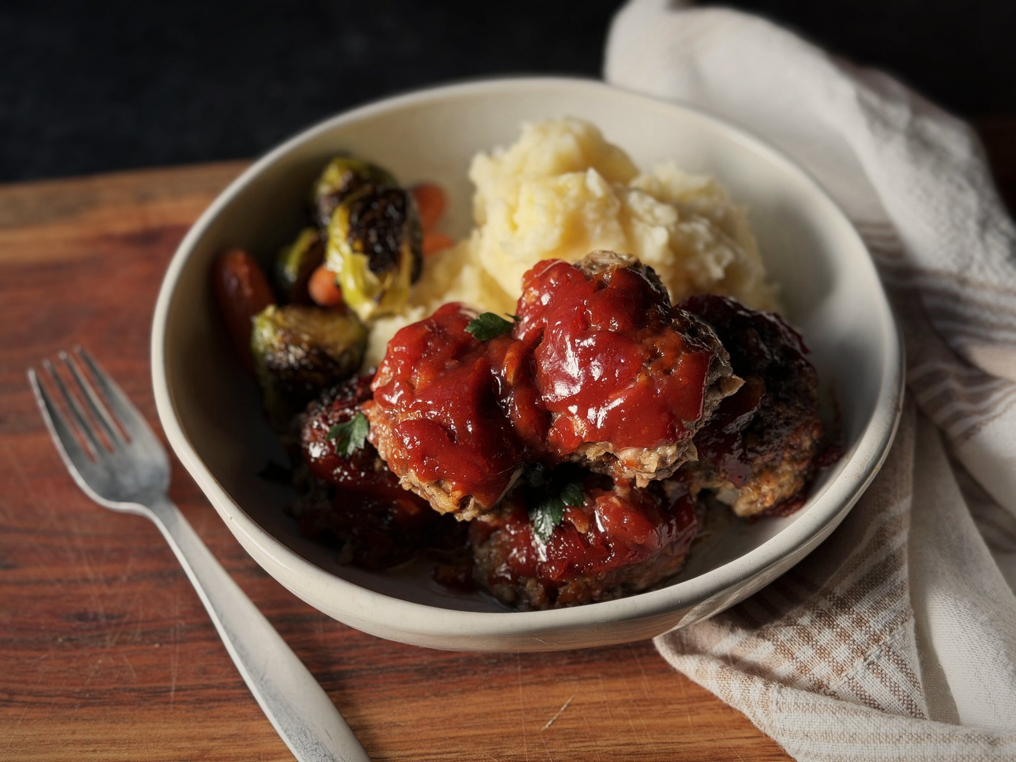Meatballs topped with red sauce and parsley in a cream bowl with mashed potatoes, roasted Brussels sprouts, and carrots, next to a fork on a wooden board.