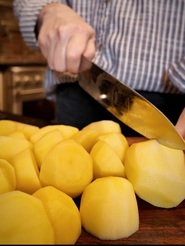 Chef's knife cutting into a peeled Yukon Gold potato beside a pile of peeled potatoes on a wooden cutting board.