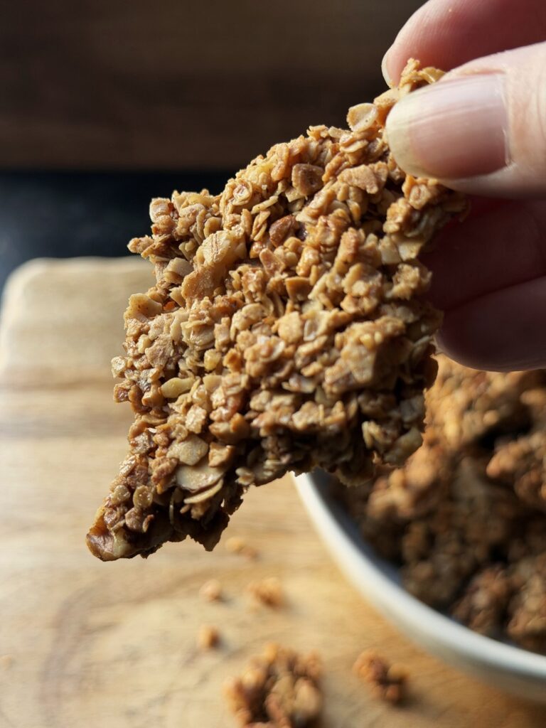 Hand holding up a large, flat granola cluster made of oats and chopped nuts above a bowl of granola.