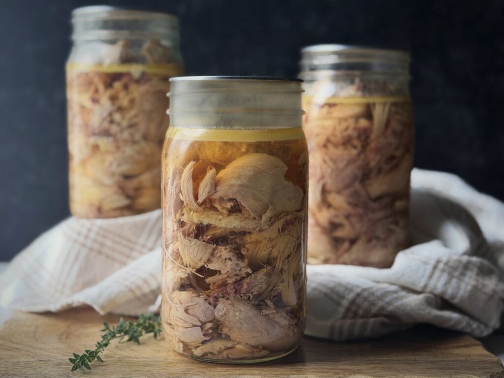 Three quart jars of home-canned chicken in golden broth on a wooden board, with a plaid linen towel and a sprig of thyme