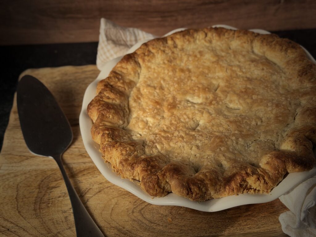 Golden all-butter pie crust with crimped, rustic edges on a chicken pot pie in a white fluted pie dish, with a pie server and linen towel on a wooden board