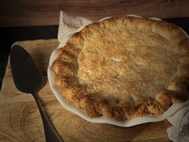 Golden all-butter pie crust with crimped, rustic edges on a chicken pot pie in a white fluted pie dish, with a pie server and linen towel on a wooden board