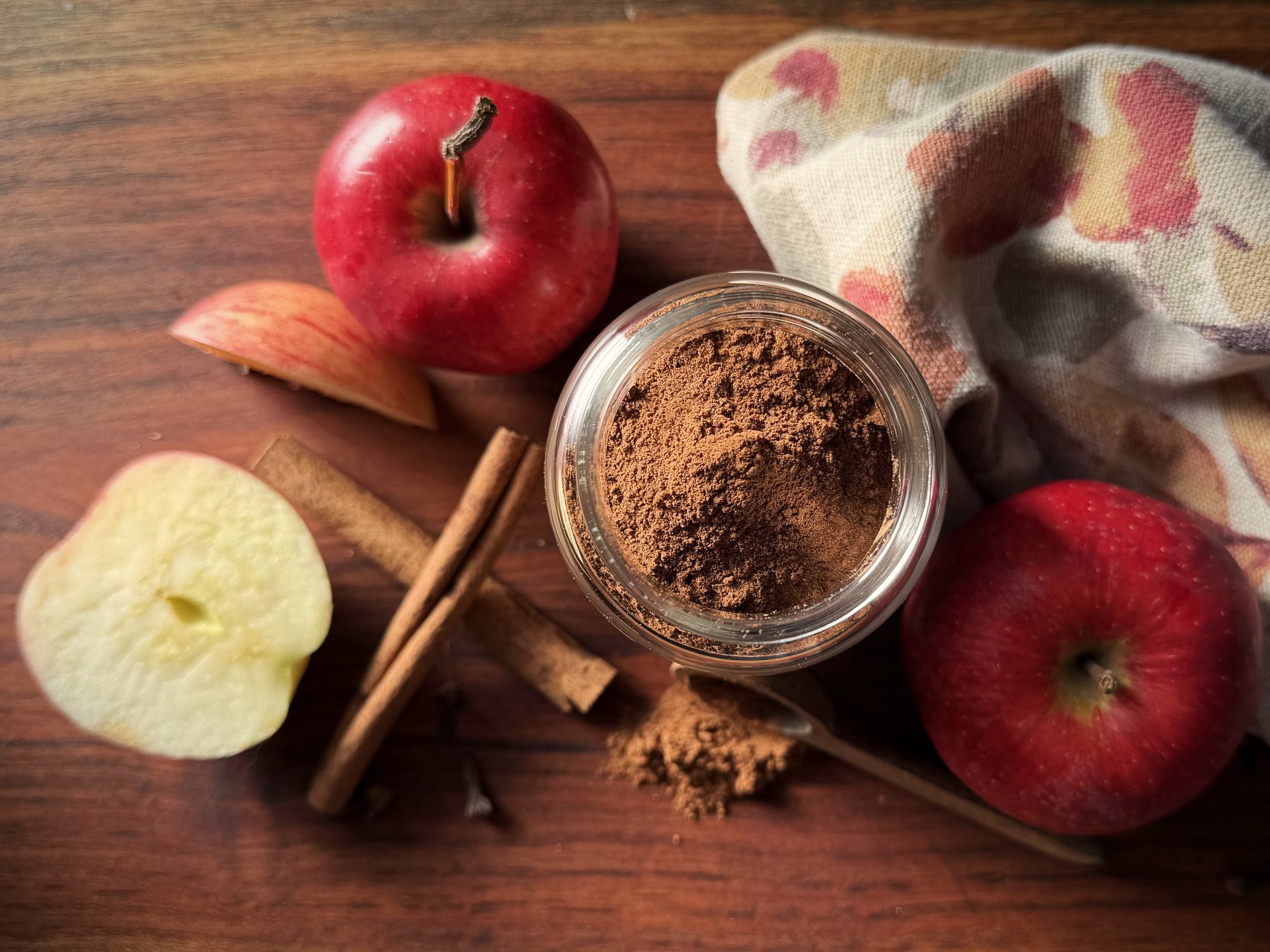 Overhead view of an open jar of homemade apple pie spice on a wooden board with red apples, apple slices, cinnamon sticks, and a floral kitchen towel