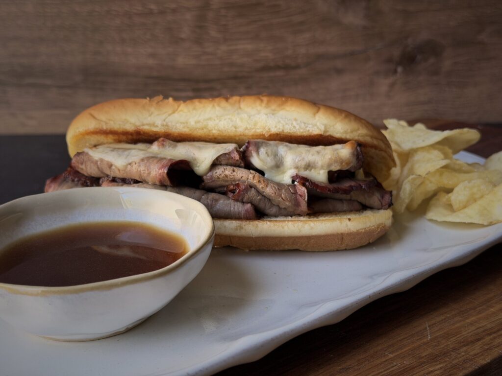 French dip sandwich with roast beef and melted provolone beside a bowl of homemade au jus and potato chips