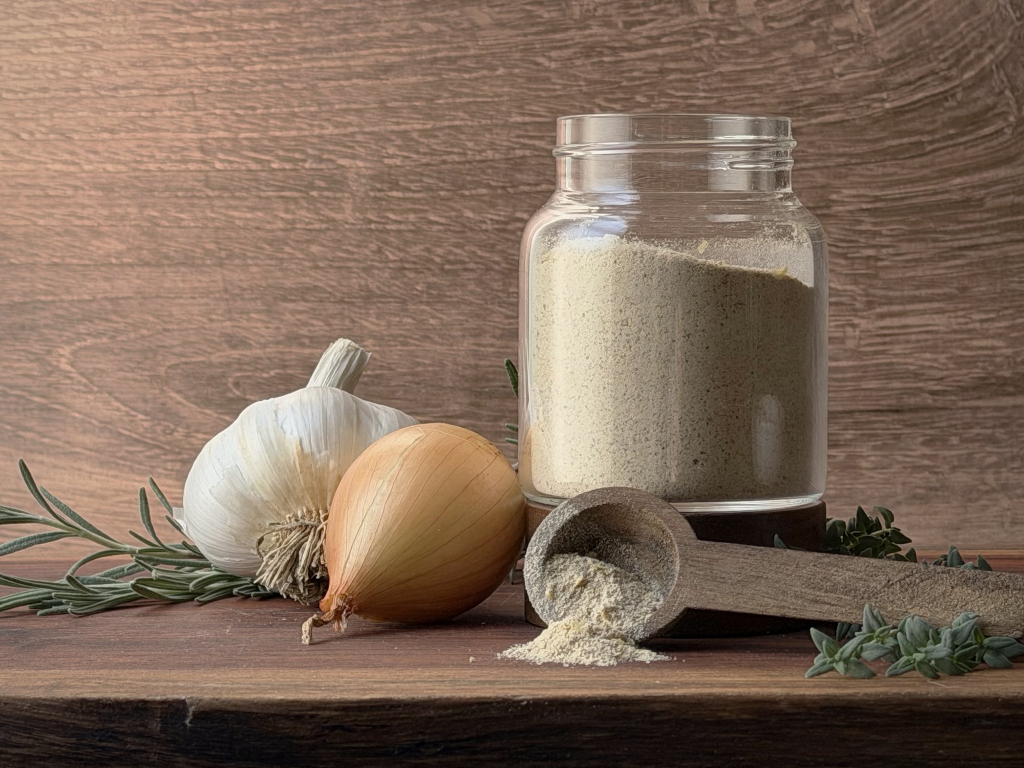 Glass jar of homemade beef bouillon powder with a wooden scoop of powder, a garlic bulb, a yellow onion, rosemary and thyme on a wooden board