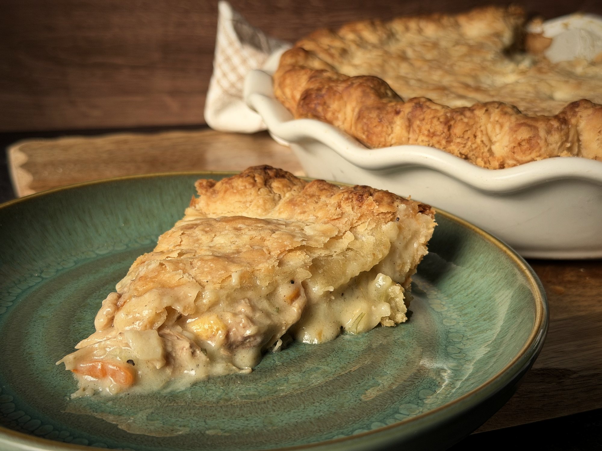 Slice of homemade chicken pot pie with creamy chicken, carrot and vegetable filling on a green plate, with the rest of the pie in a white fluted dish behind it