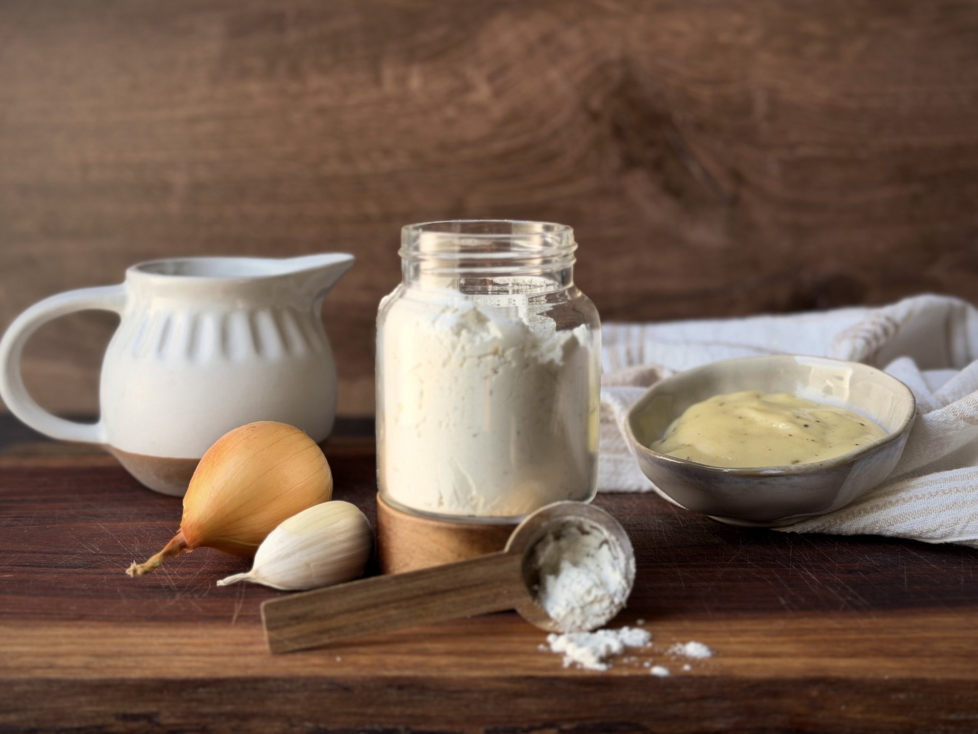 Glass jar of dry cream of soup mix with a wooden scoop, a bowl of cream soup, onion, garlic and a white pitcher on a wooden board