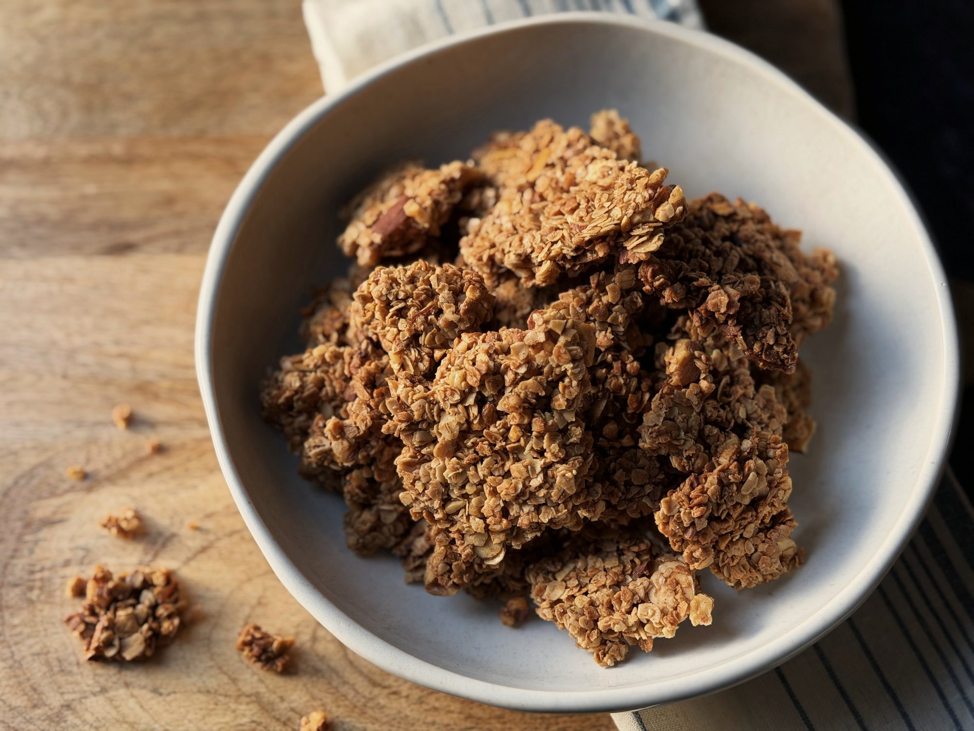 Large homemade granola clusters piled in a white bowl on a wooden board, with a few loose pieces and a striped towel beside it.