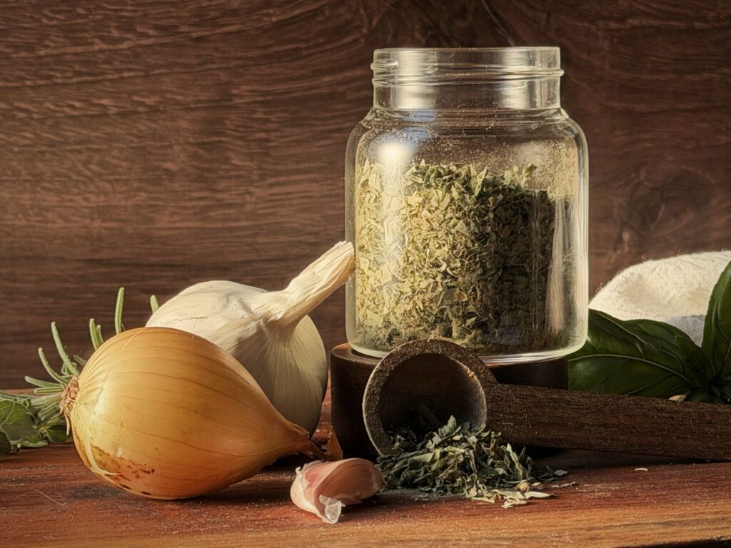 Glass jar of homemade Italian seasoning with a wooden scoop of dried herbs, an onion, garlic, fresh basil and rosemary on a wooden board