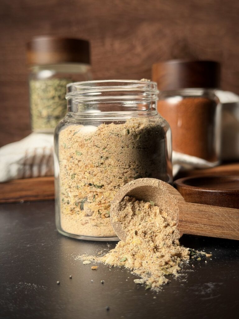 Glass jar of homemade onion soup mix with a wooden scoop spilling mix onto a dark counter