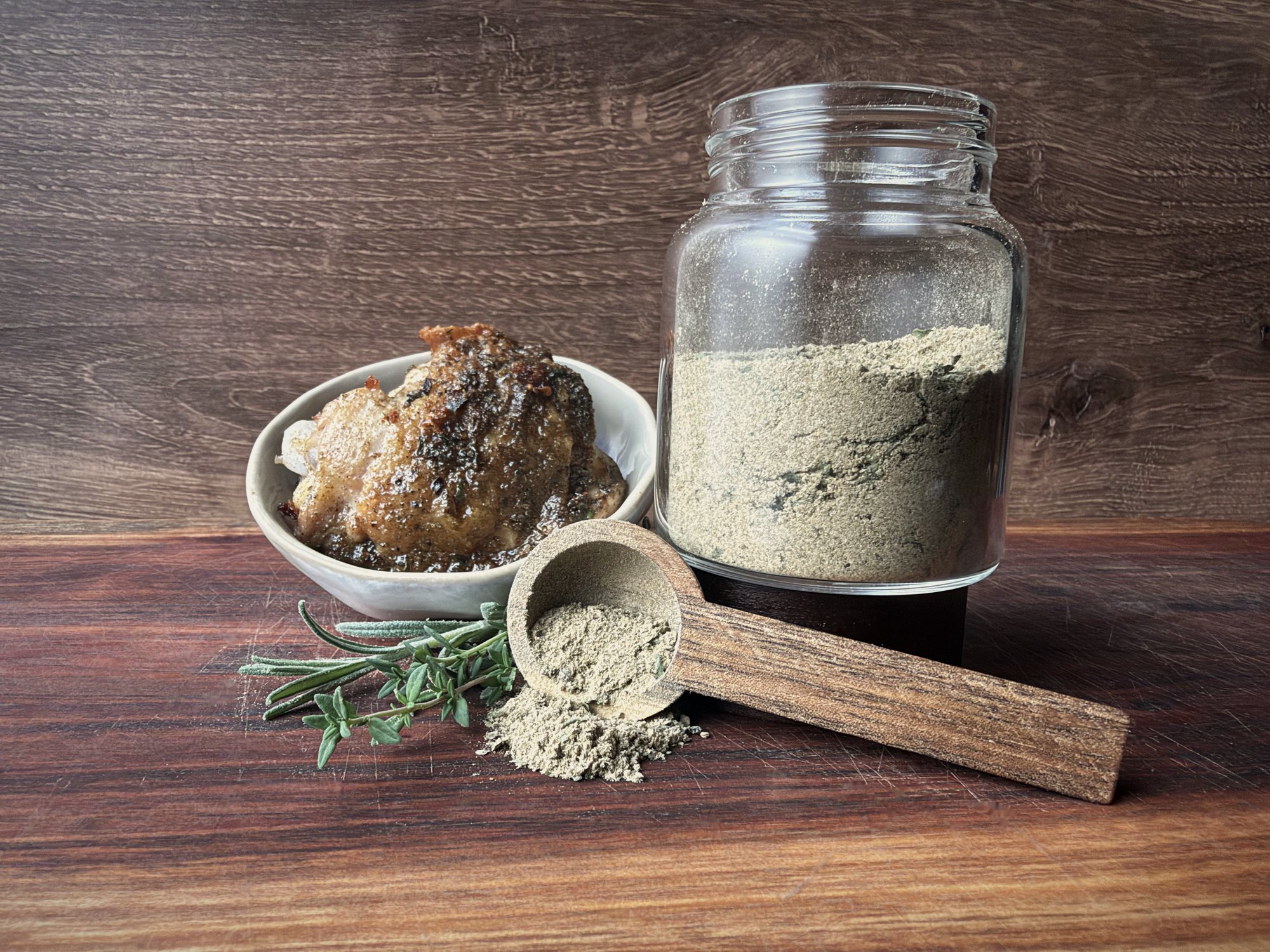 Glass jar of homemade poultry seasoning beside a bowl of browned chicken, with a wooden scoop of seasoning and sprigs of rosemary and thyme on a wood board