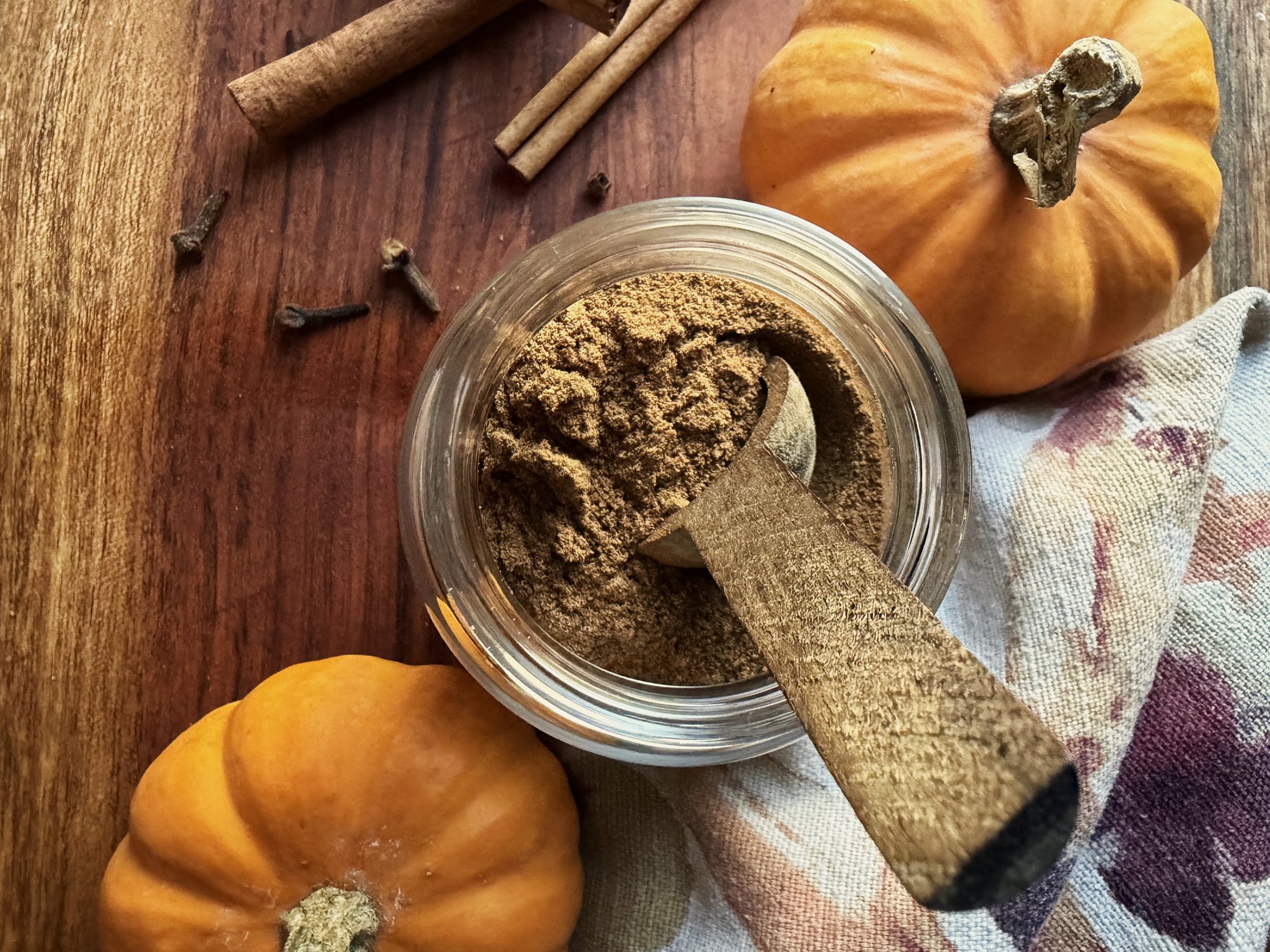 Glass jar of homemade pumpkin pie spice with a wooden scoop, surrounded by mini pumpkins, cinnamon sticks, whole cloves and a floral linen towel on a wood board