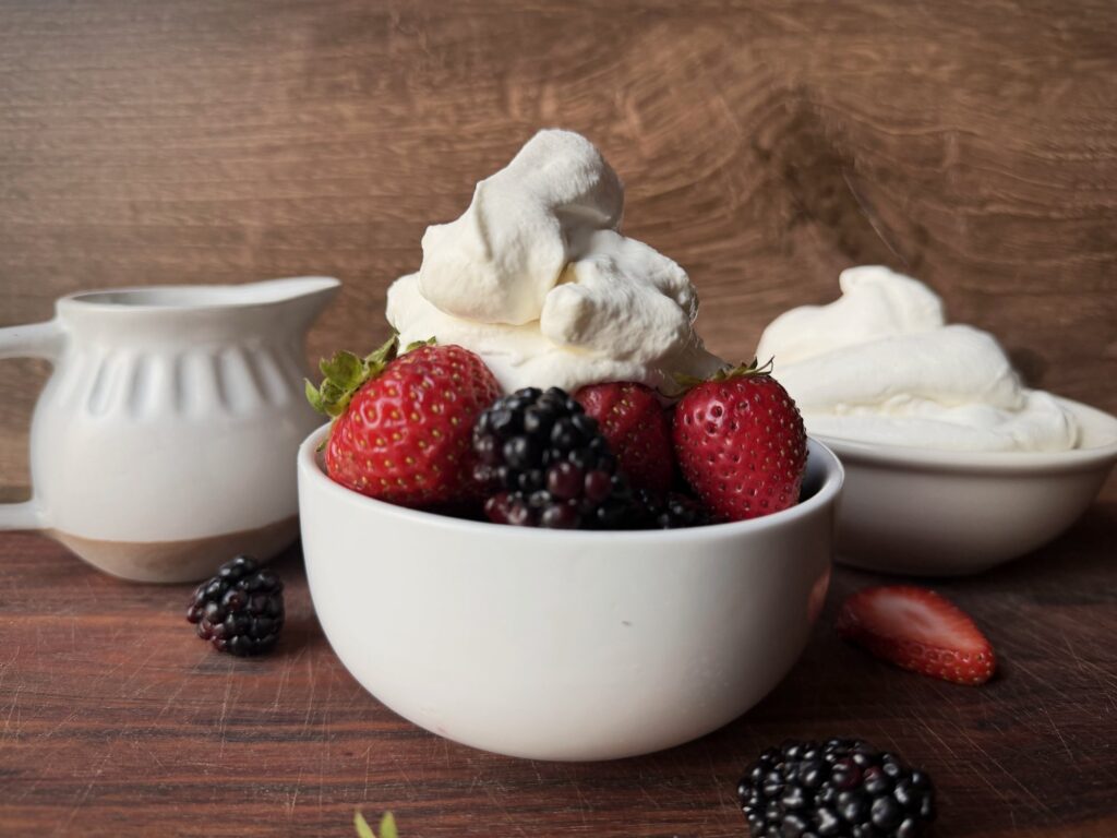 White bowl of strawberries and blackberries topped with a tall swirl of homemade whipped cream, with a cream pitcher and a bowl of whipped cream behind it