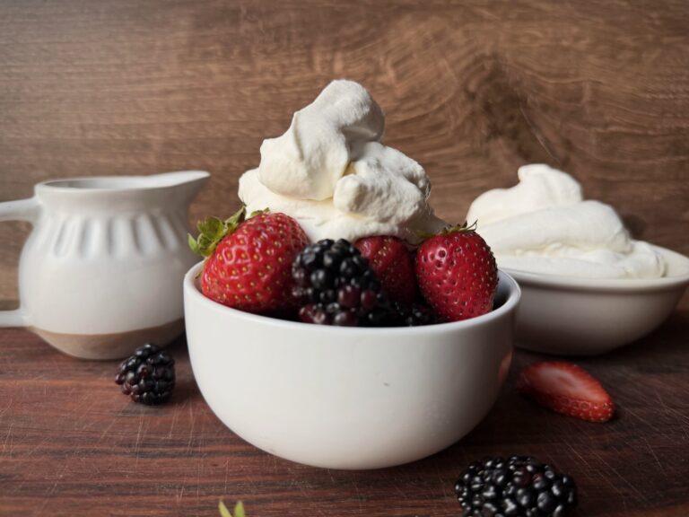 White bowl of strawberries and blackberries topped with a tall swirl of homemade whipped cream, with a cream pitcher and a bowl of whipped cream behind it