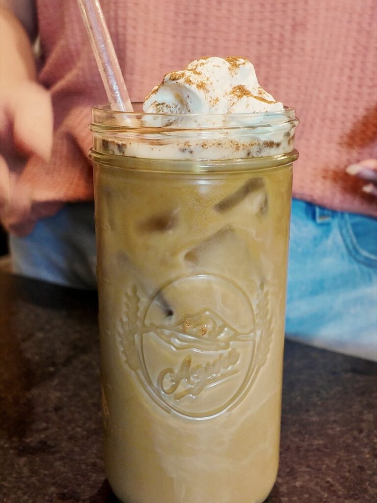 Iced coffee drink in a tall glass jar topped with whipped cream and a sprinkle of pumpkin pie spice, with a glass straw