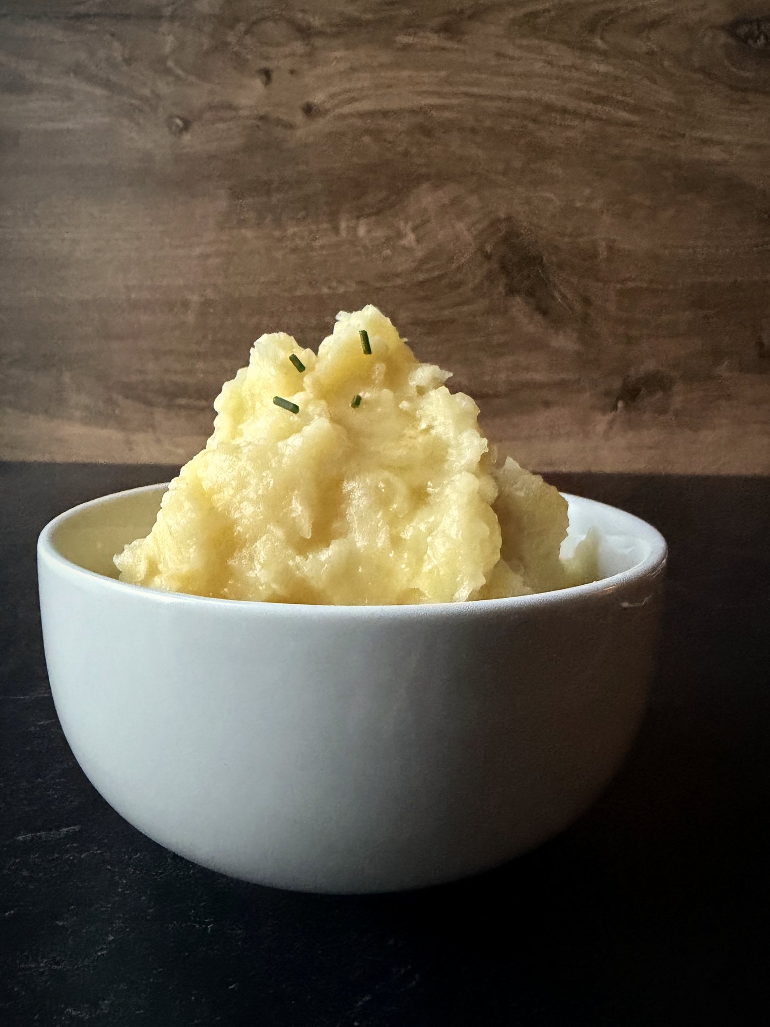Yukon Gold mashed potatoes piled high in a white bowl, sprinkled with a few chives, on a dark countertop in front of a wood backdrop.
