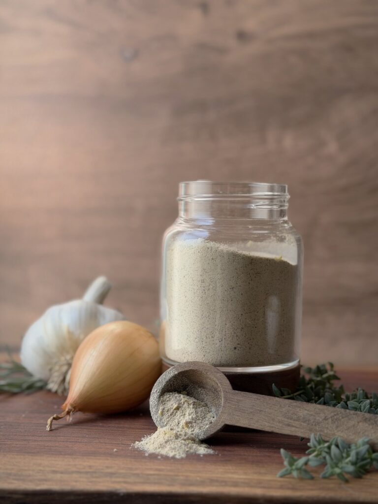 Jar of beef bouillon powder beside a garlic bulb, yellow onion and wooden scoop