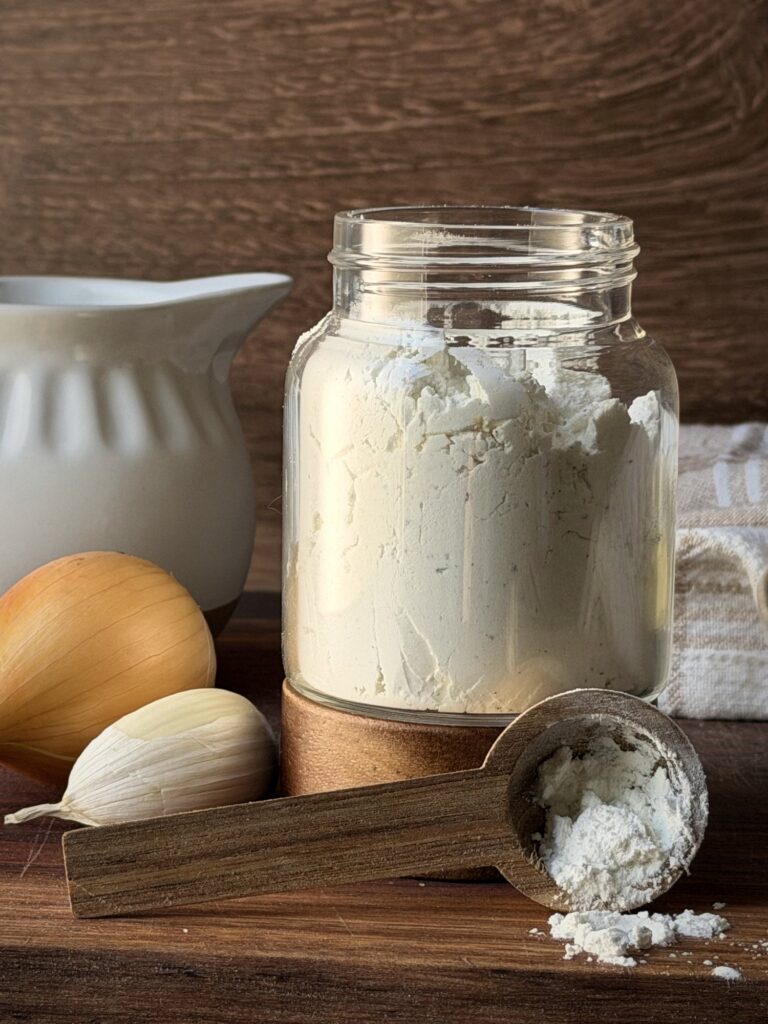 Close-up of a glass jar of dry cream of soup mix with a wooden scoop, onion and garlic