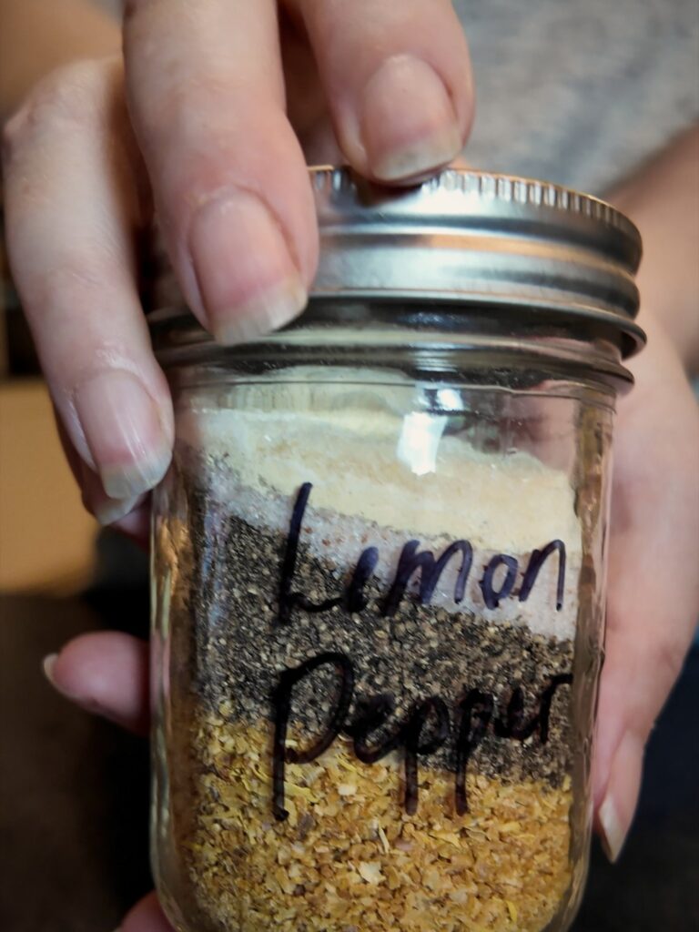 Hand holding a jar of layered lemon pepper seasoning labeled Lemon Pepper