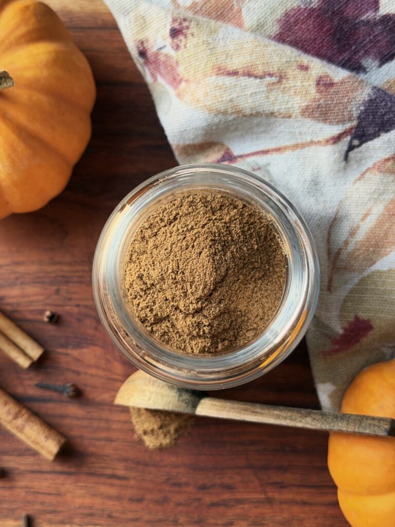 Overhead view of an open jar of homemade pumpkin pie spice next to a wooden scoop of spice, mini pumpkins, cinnamon sticks and cloves