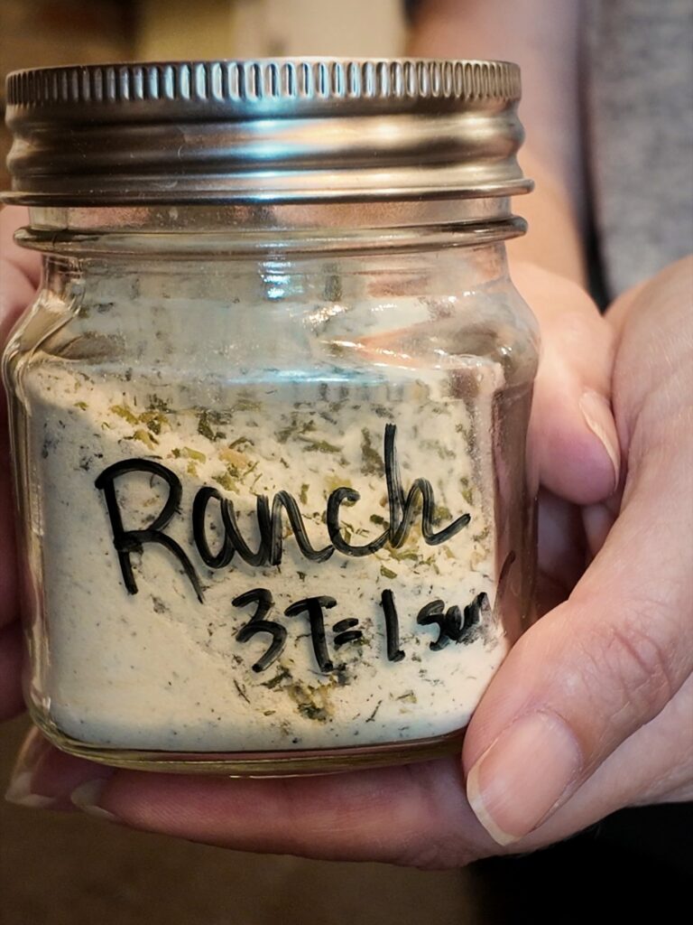 Hand holding a jar of homemade ranch seasoning mix labeled "Ranch 3T = 1 pkg"