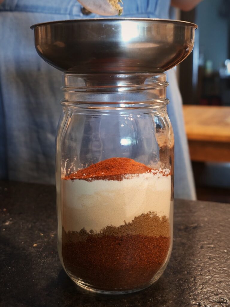 Spooning spices through a funnel into a jar layered with fajita seasoning ingredients