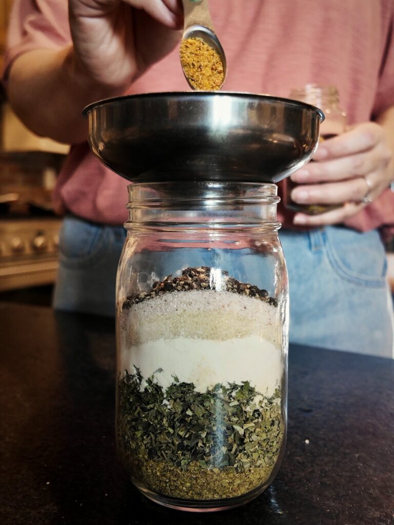Spooning seasoning through a funnel into a mason jar layered with dried herbs and powders