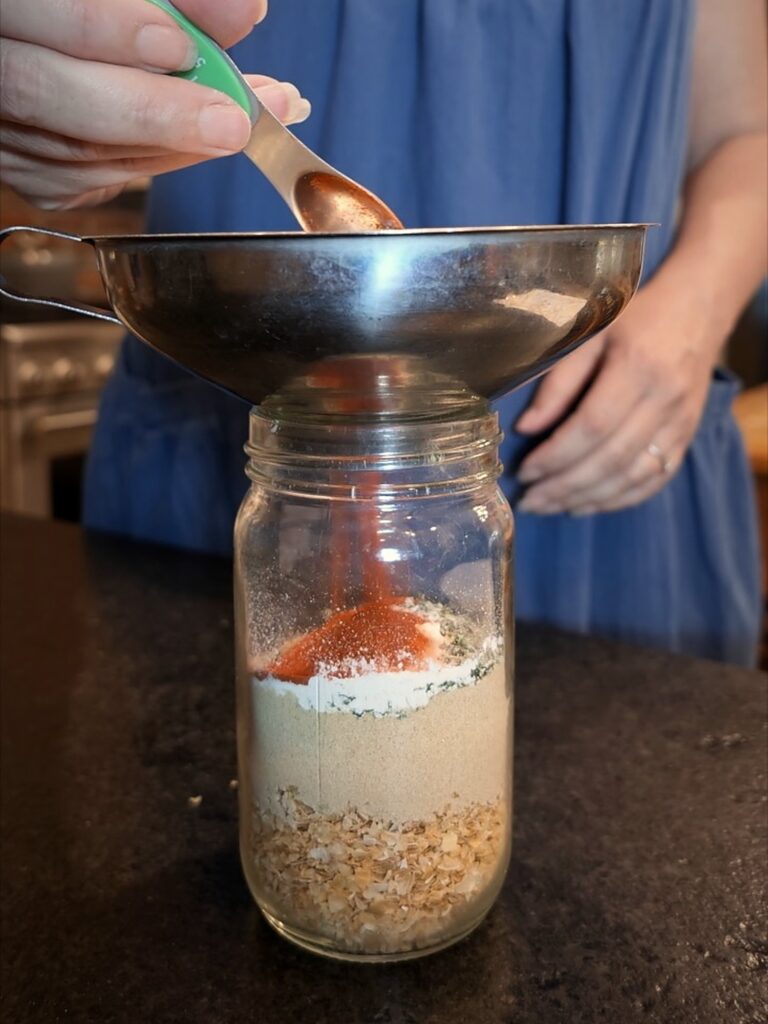 Measuring spoon adding red seasoning through a canning funnel into a jar of layered onion soup mix ingredients