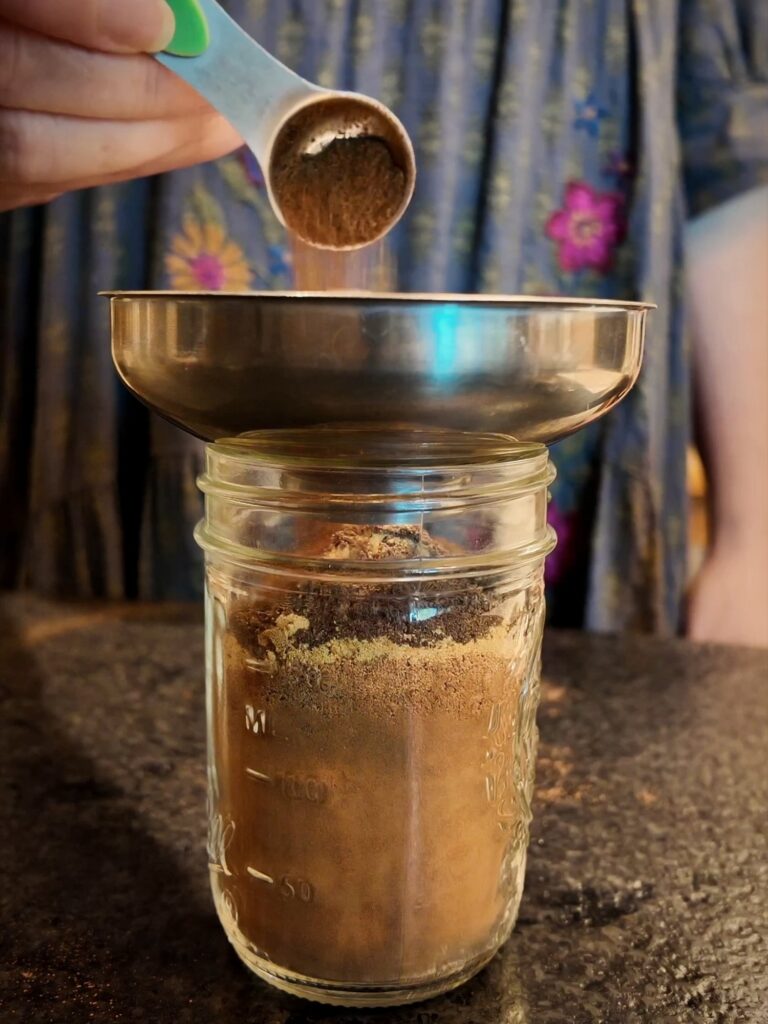 Measuring spoon of ground spice over a funnel on a glass jar with layers of ground spices