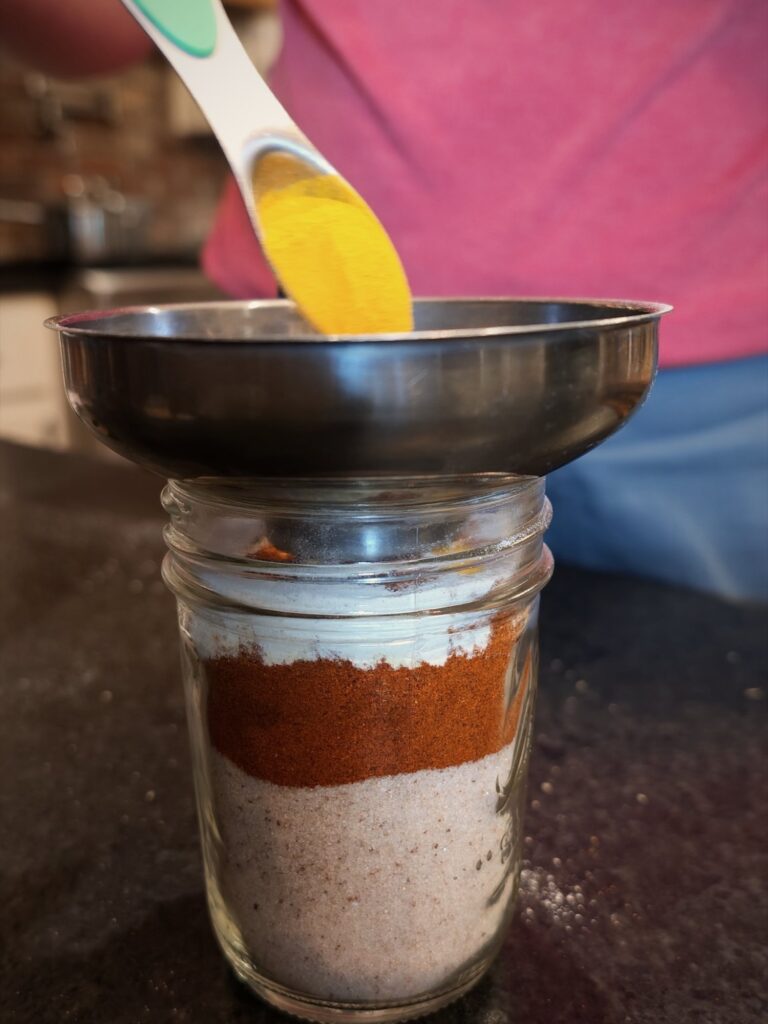 Spooning turmeric through a funnel into a jar layered with salt and paprika