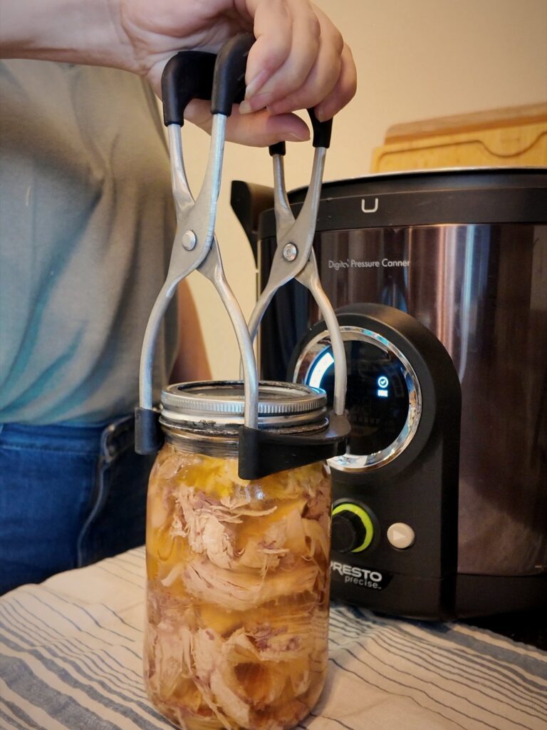 Jar lifter setting a hot jar of canned chicken onto a striped towel in front of a Presto digital pressure canner