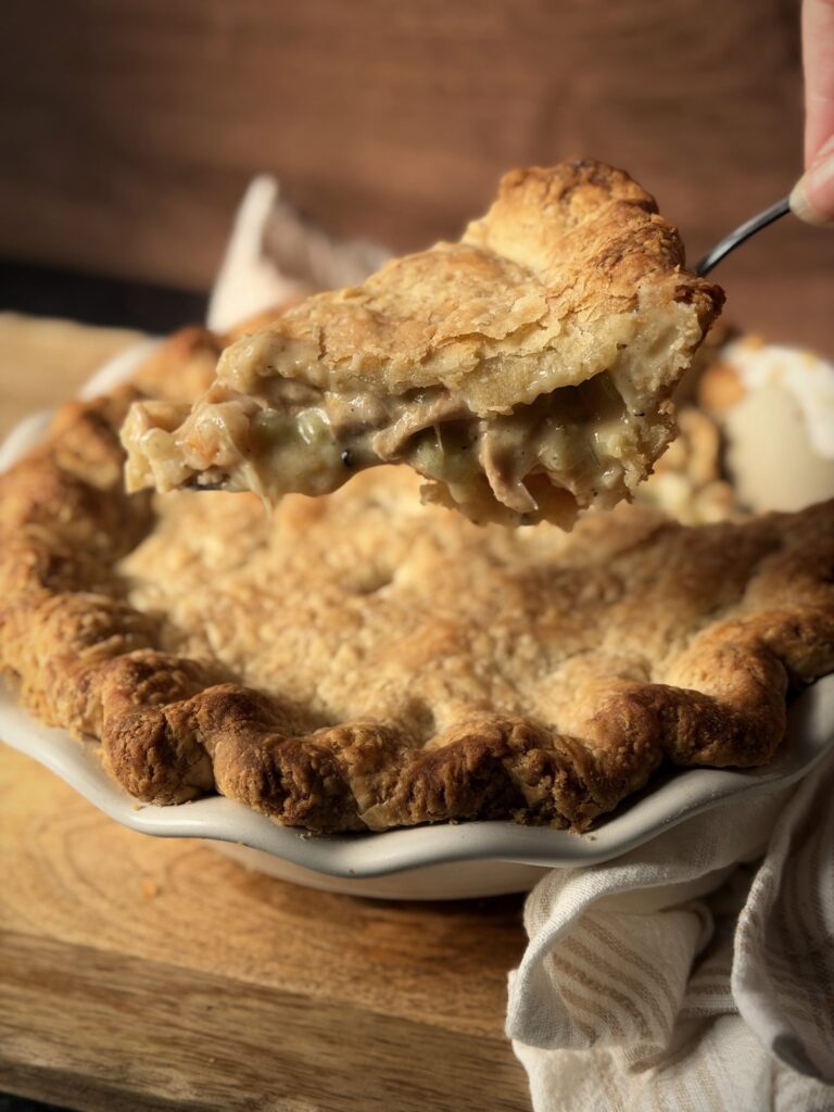 Server lifting a slice of chicken pot pie out of a white fluted pie dish, showing the creamy filling under the flaky crust