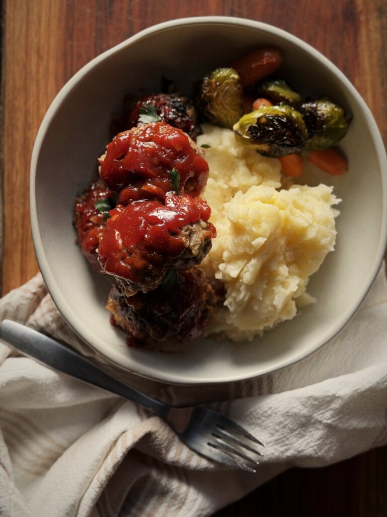 Overhead view of sauced meatballs with mashed potatoes, roasted Brussels sprouts, and carrots in a cream bowl, with a fork and striped towel.