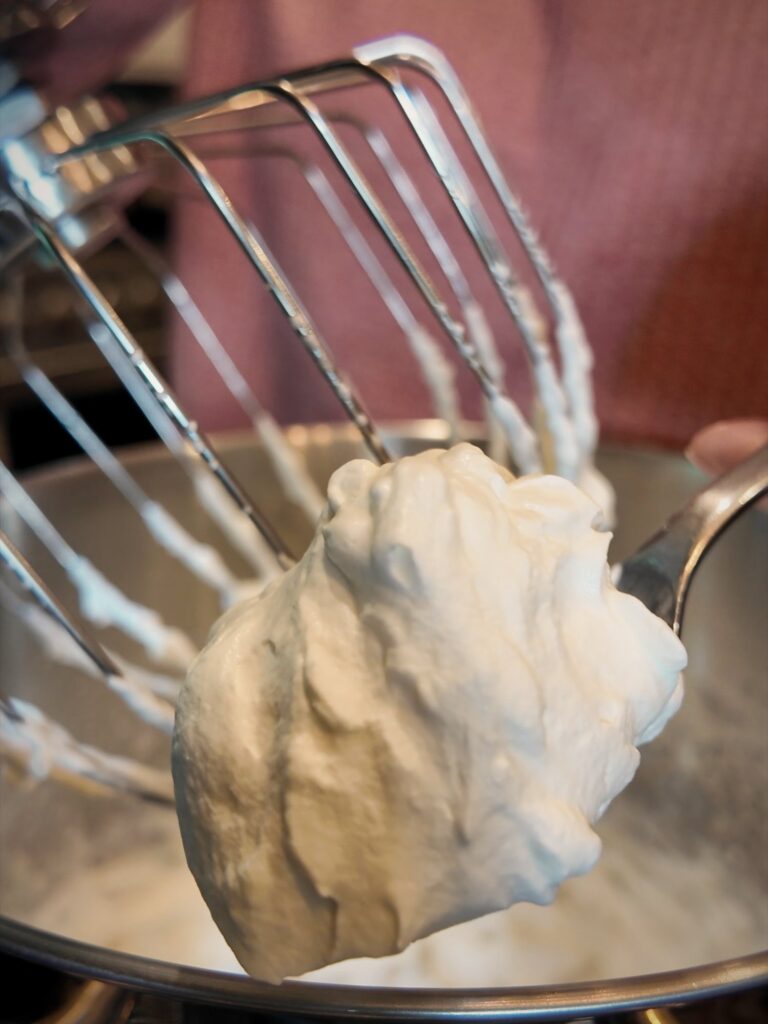 Thick mound of whipped cream clinging to a stand mixer whisk above the bowl