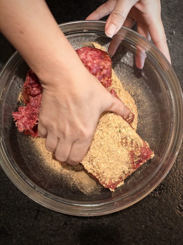 Hands mixing onion soup mix into ground beef in a glass bowl