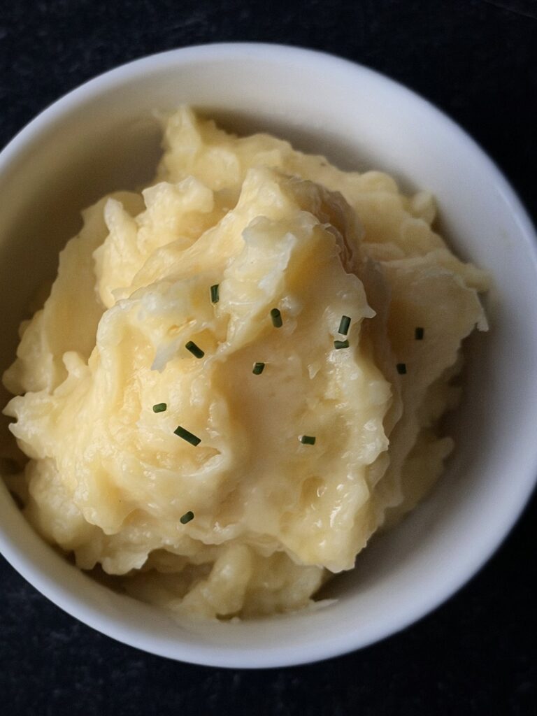 Overhead view of creamy Yukon Gold mashed potatoes in a white bowl with a few chives on top.