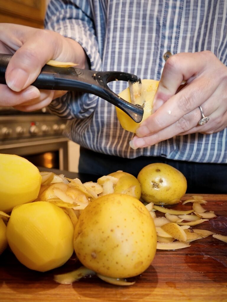 Hands peeling a Yukon Gold potato with a vegetable peeler over a cutting board with peels and whole potatoes.