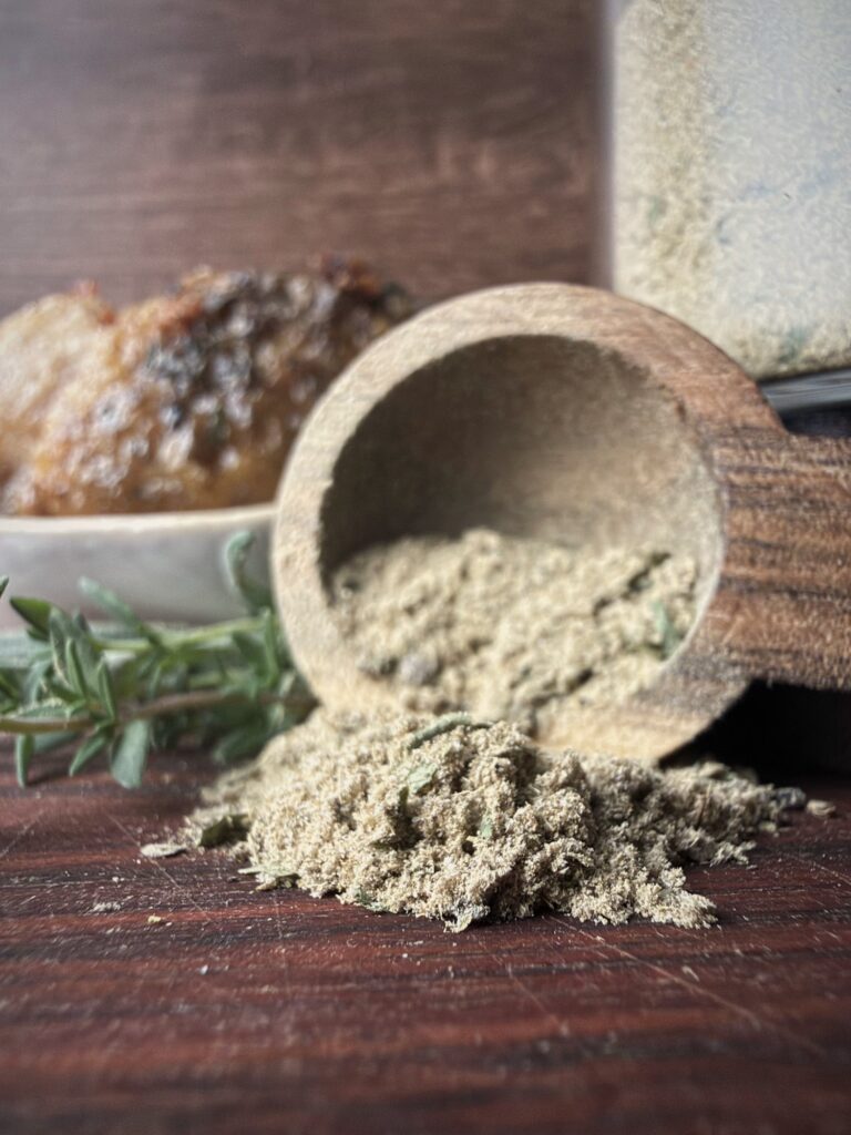 Wooden scoop tipped over with poultry seasoning spilling onto a wood board, with rosemary and chicken in the background