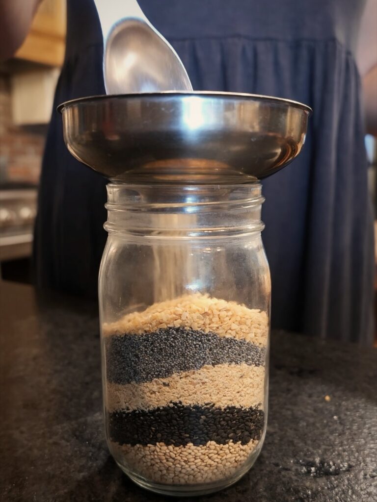 Measuring spoon over a stainless steel funnel on a mason jar layered with seeds and dried onion