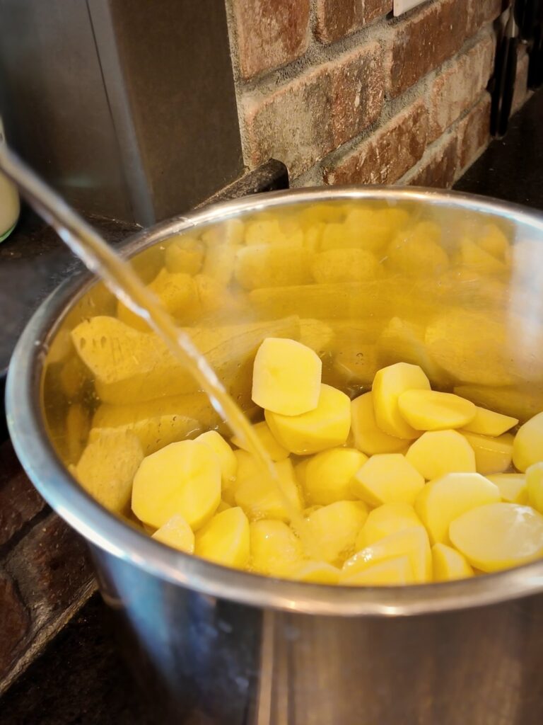 Water pouring from a faucet into a stainless steel pot of chopped, peeled Yukon Gold potatoes.
