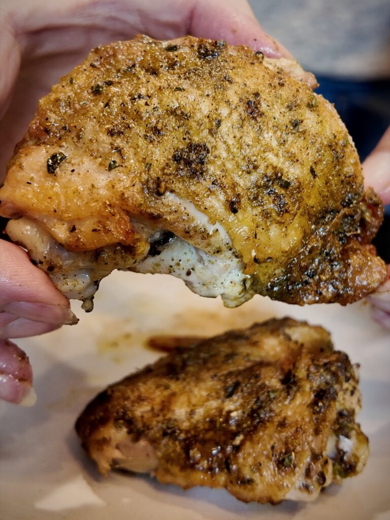 Hand holding a seared chicken thigh coated in herb seasoning above a second piece on a plate