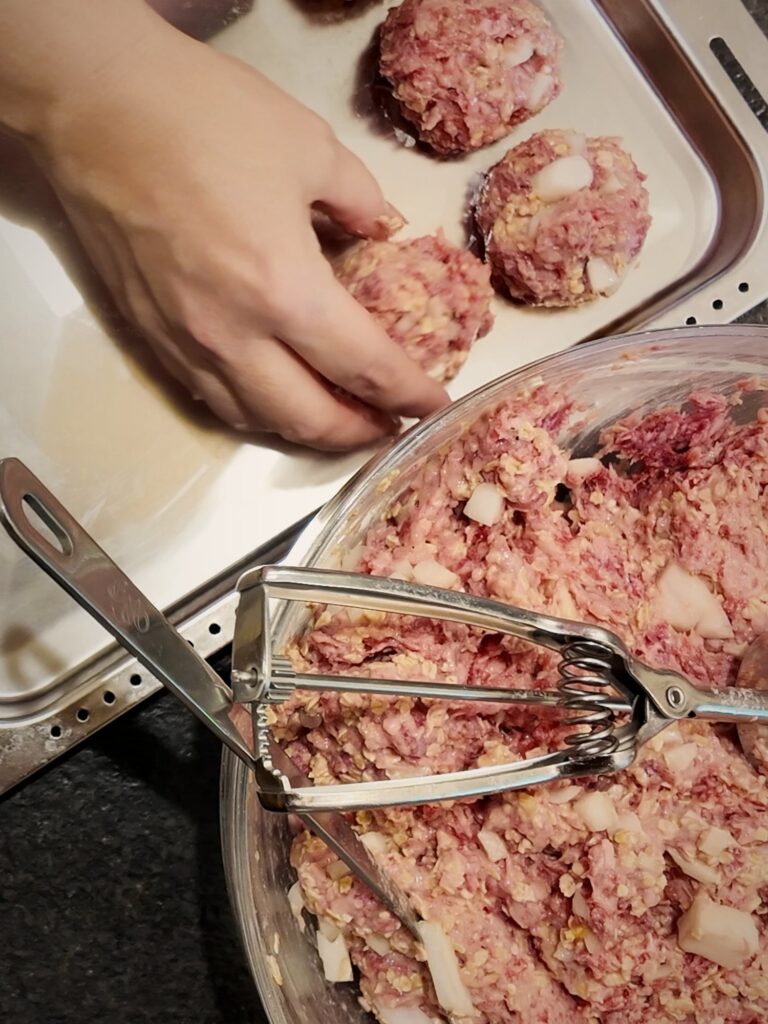 Hand placing scooped meatballs in a metal baking pan beside a bowl of meatball mixture and a cookie scoop.