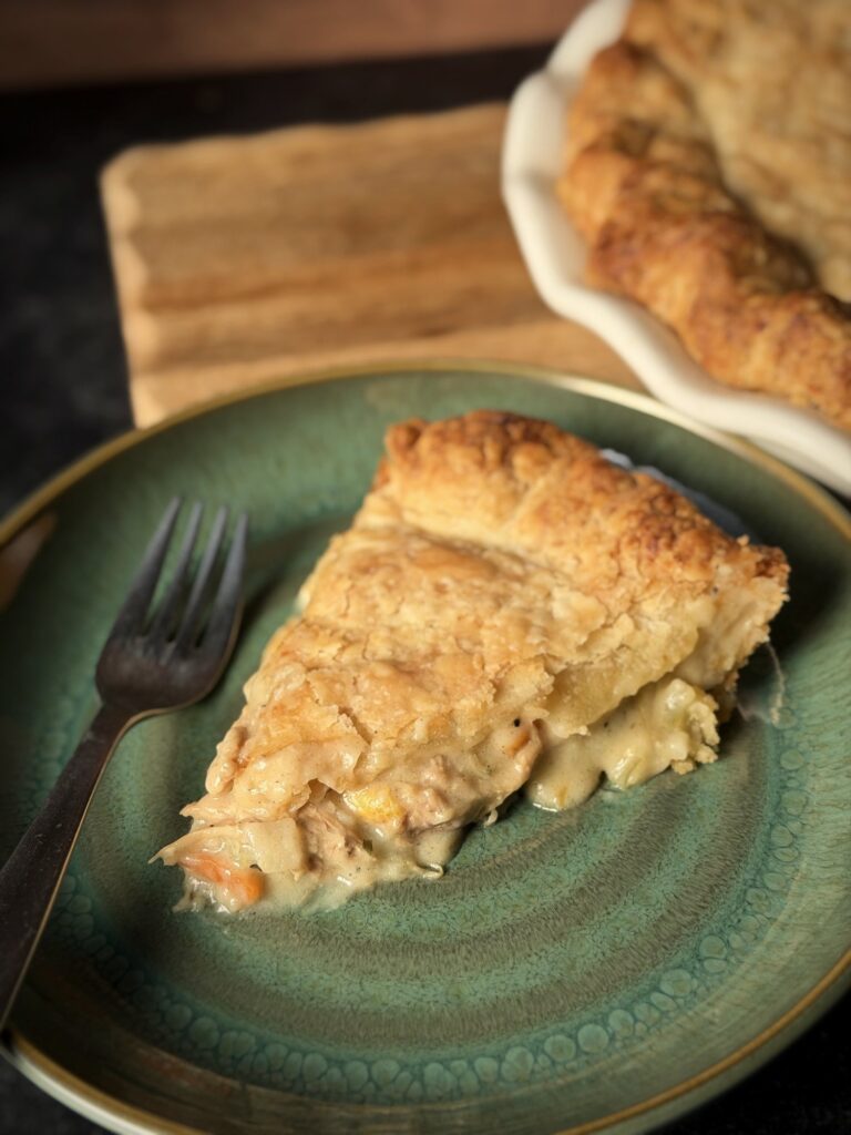 Slice of chicken pot pie on a green plate with a fork, with the baked pie in the background on a wooden board