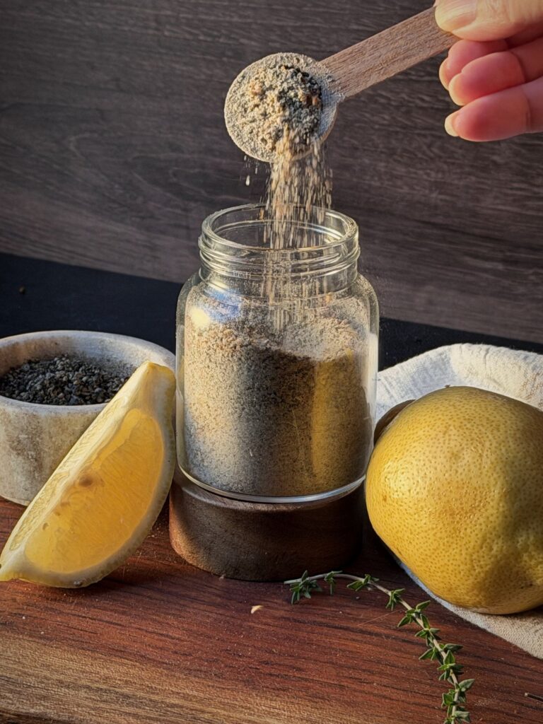 Pouring lemon pepper seasoning from a wooden scoop into a glass jar