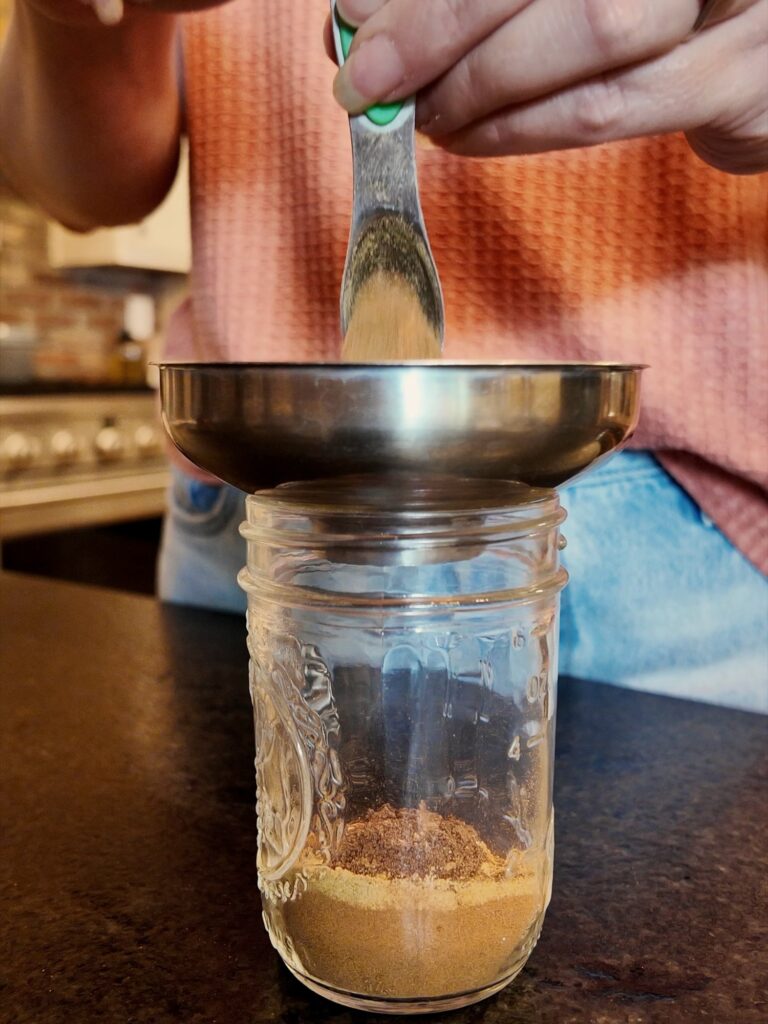 Measuring spoon adding ground spice through a stainless steel funnel into a small glass jar