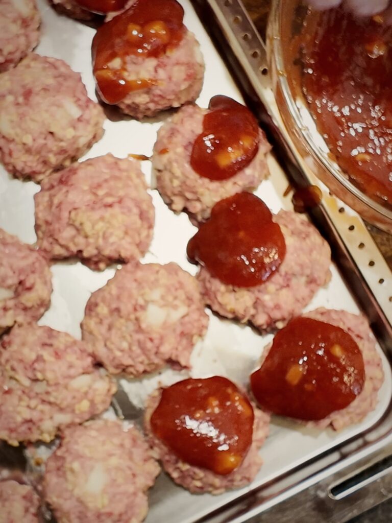 Spooning red sauce over raw meatballs arranged in a single layer in a metal baking pan.