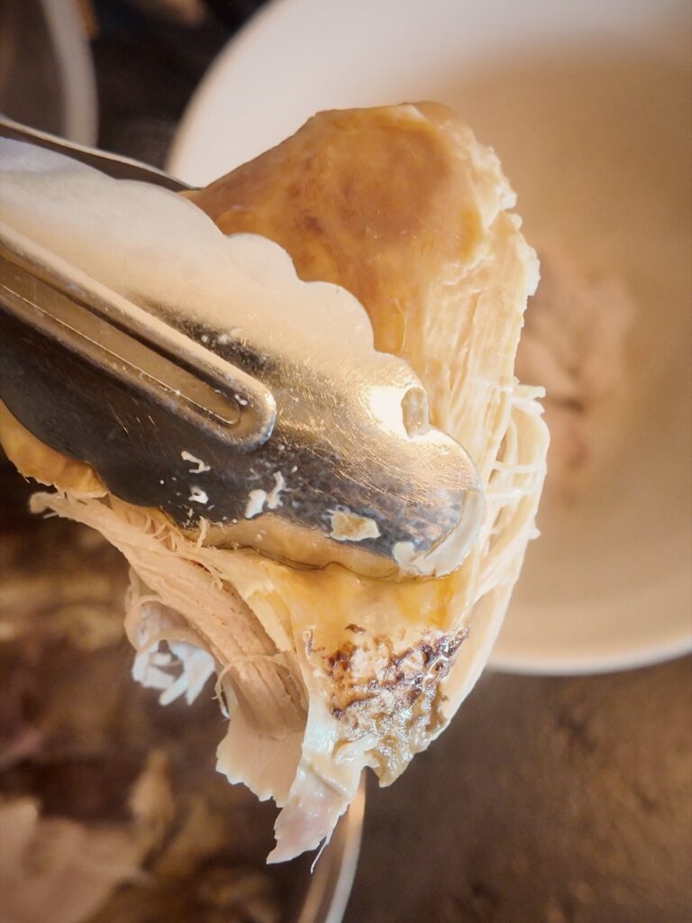 Tongs lifting a large, tender piece of cooked chicken over a white bowl