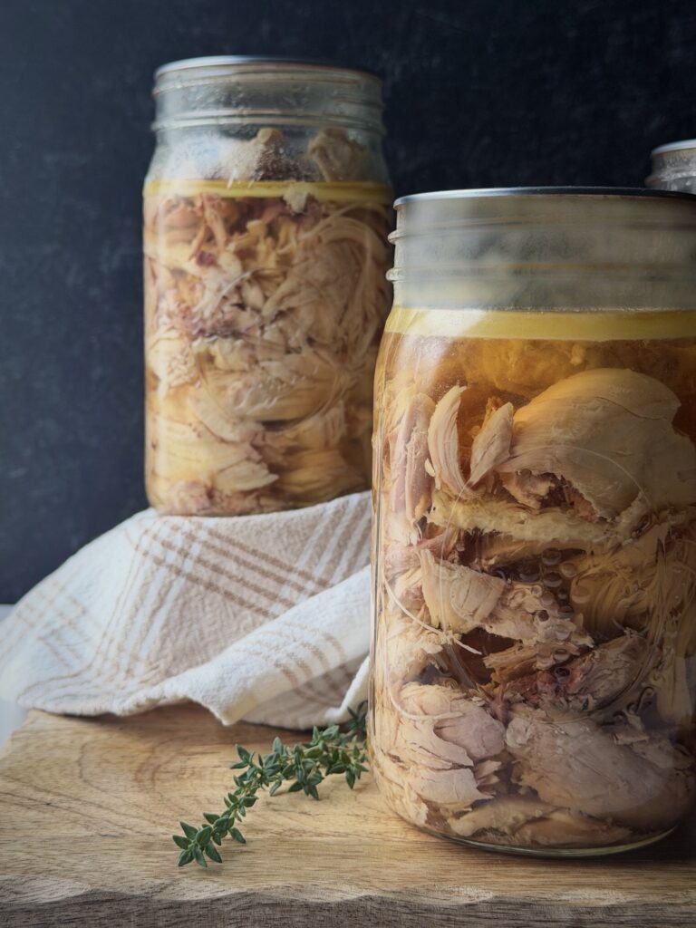 Two quart jars of home-canned chicken in broth on a wooden board with a plaid linen towel and a sprig of thyme