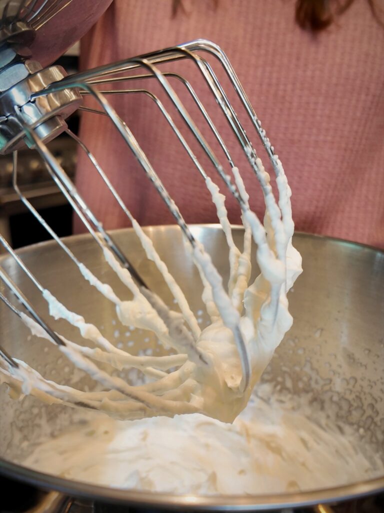 Stand mixer whisk coated in whipped cream above a bowl of whipped cream