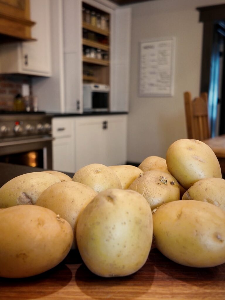 A pile of whole, unpeeled Yukon Gold potatoes on a wooden cutting board in a kitchen.